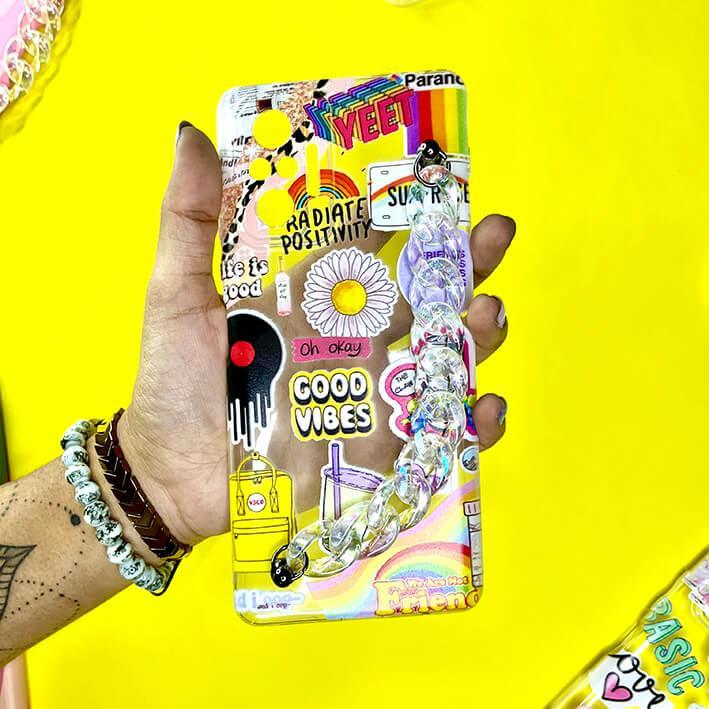 Colorful phone case with stickers, chain handle, and positive vibes design held on yellow background