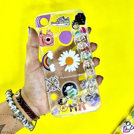 Clear phone case with colorful stickers and chain charm, held in hand on yellow background