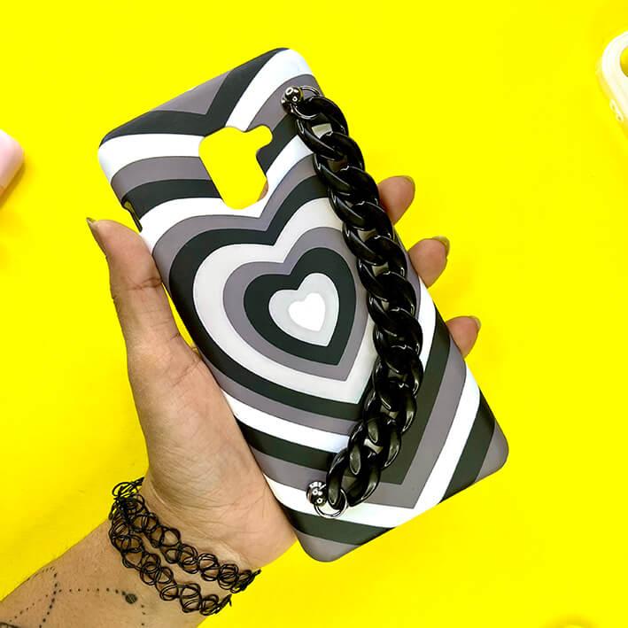 Black and white heart pattern phone case with black chain charm, held in hand on yellow background.