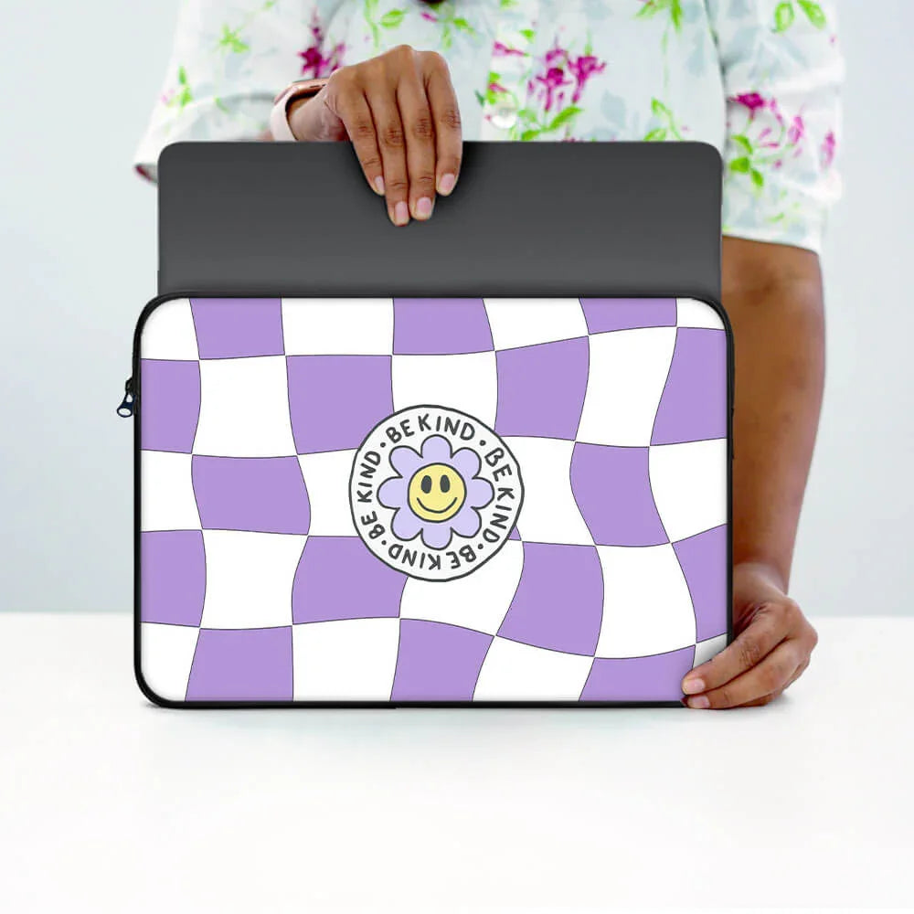 Person holding a laptop sleeve with purple and white checkered pattern, smiley flower, and 'Be Kind' text.