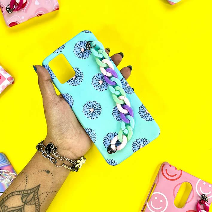 Hand holding blue floral phone case with pastel chain charm on yellow background