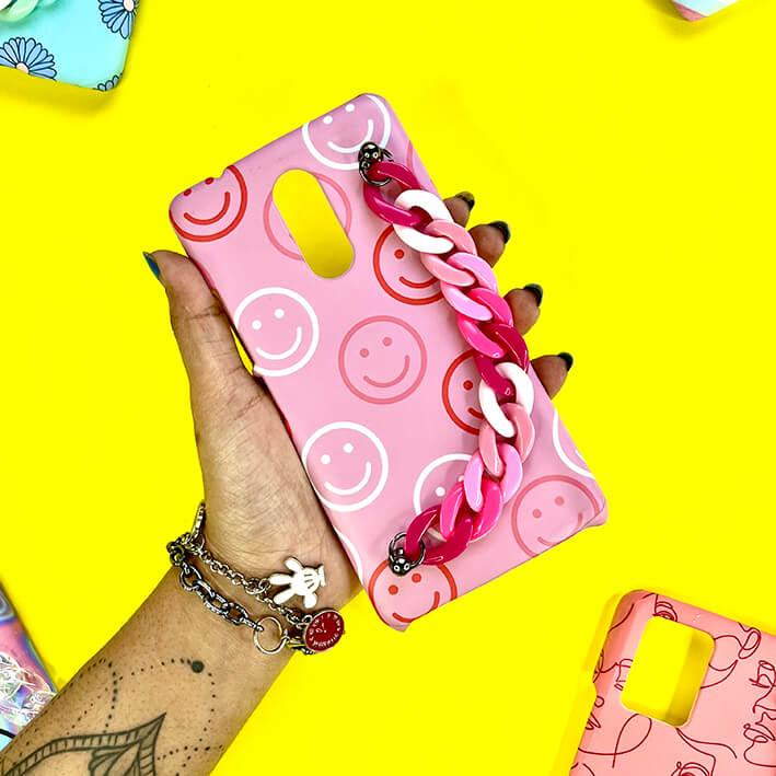 Pink smiley face phone case with chunky pink chain strap, held in hand on yellow background